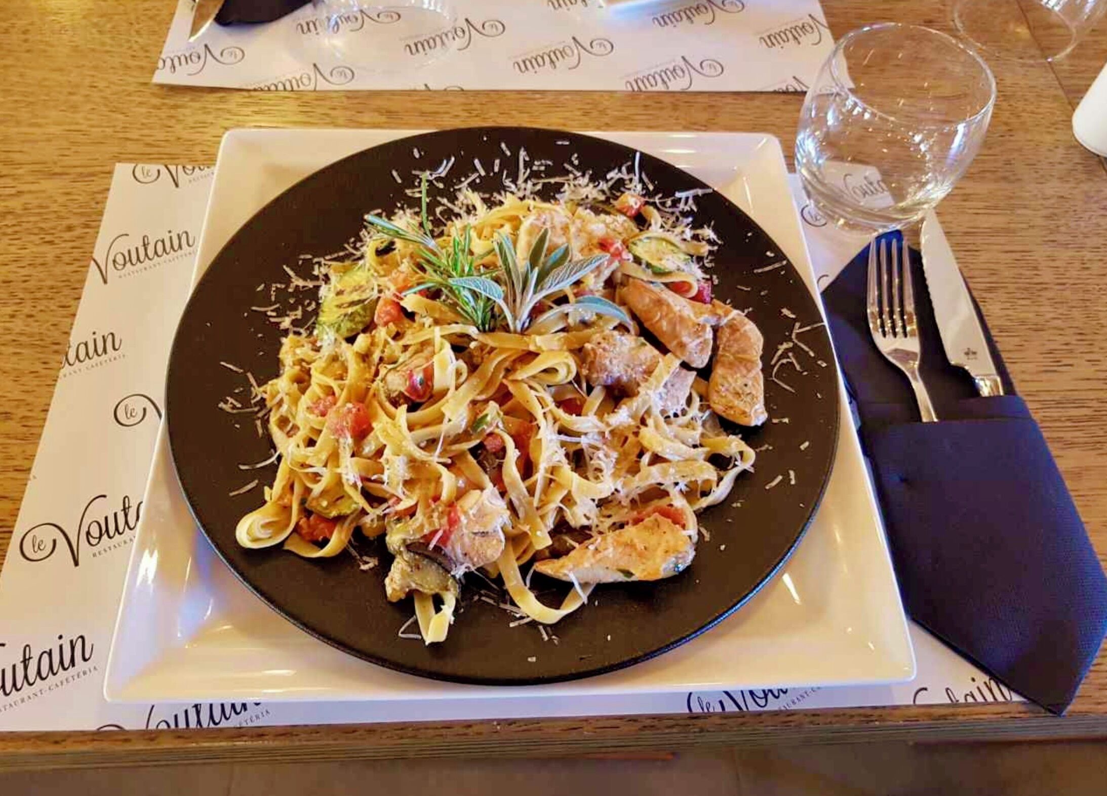A plate of pasta with chicken and vegetables on a black plate, placed on a white tablecloth with a napkin and fork to the side.