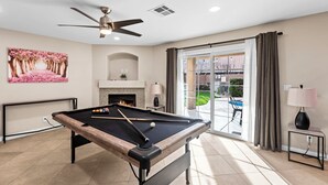 Une table de billard dans une pièce dotée d’un ventilateur de plafond, d’une cheminée et donnant sur une terrasse aménagée avec du mobilier d’extérieur.