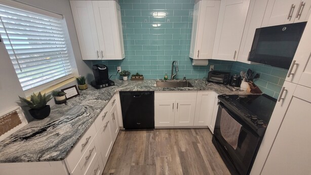 A kitchen with white cabinets, a black dishwasher, a sink with a faucet, and a countertop with a coffee maker and plants. - Cozy Cottage Sleeps 8 close to the World Equestrian Center (Ocala)