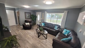 A living room with a leather sofa, a leather armchair, and a glass coffee table. There is a window with curtains, a door, and a framed picture on the wall. - Cozy Cottage Sleeps 8 close to the World Equestrian Center (Ocala)
