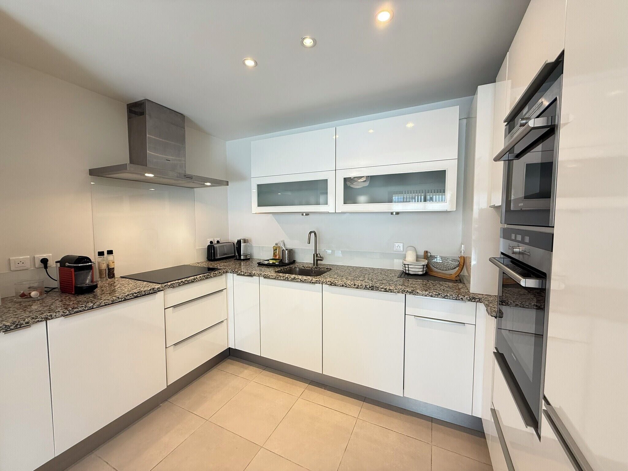 A modern kitchen with white cabinets, granite countertops, a built-in oven, and a stainless steel range hood.
