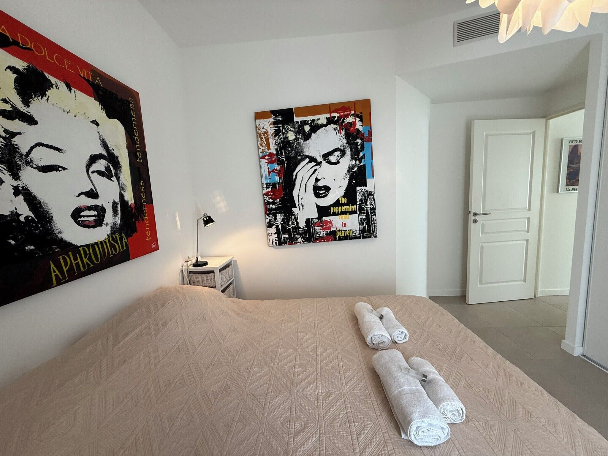 A bedroom with a bed, two rolled towels, a nightstand with a lamp, and two wall art pieces.