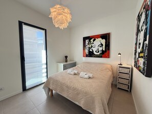 Une chambre à coucher comprenant un lit, une table de chevet, une lampe, une fenêtre avec des stores et un mur décoré d’œuvres d’art.
