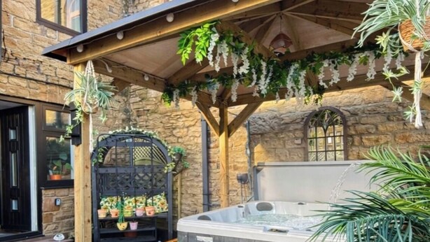 Een achtertuin met een jacuzzi, een pergola met hangplanten, een stenen muur en een houten terras.