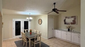 A dining area with a table and chairs, a ceiling fan, a clock, and a kitchen with cabinets and a countertop. - Relaxed Desert Stay - 3BR/2BA Tucson Home - Furnished & Spacious (Tucson)