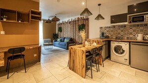 Een kleine keuken met een wasmachine, een eethoek met een houten tafel, en een bank op de achtergrond.