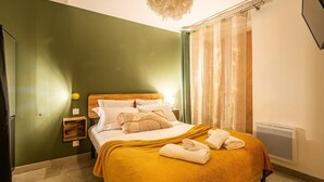 Een net opgemaakt bed met een gele deken, witte kussens en een houten hoofdbord. Er staat een klein nachtkastje met een lamp en een wandplank.