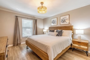 A bedroom with a wooden bed, a nightstand with a lamp, a window with curtains, and two framed pictures on the wall. - Beach Tyme Retreat - Park Shore Resort Near Beach (Naples)