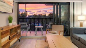 En balkon med udsigt til solnedgang, et træbord, to stole, en sofa og et fjernsyn monteret på væggen.