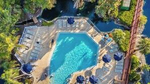 Oversigtsbillede af et resort med en stor, rektangulær pool omgivet af liggestole og parasoller, foran frodigt grønt område og en klippefyldt kystlinje.