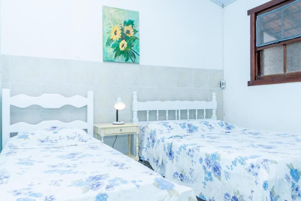 Une chambre avec deux lits simples, une table de chevet avec une lampe, un tableau représentant des fleurs accroché au mur et une fenêtre avec un cadre en bois.