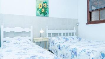 Une chambre avec deux lits simples, une table de chevet avec une lampe, un tableau représentant des fleurs accroché au mur et une fenêtre avec un cadre en bois.