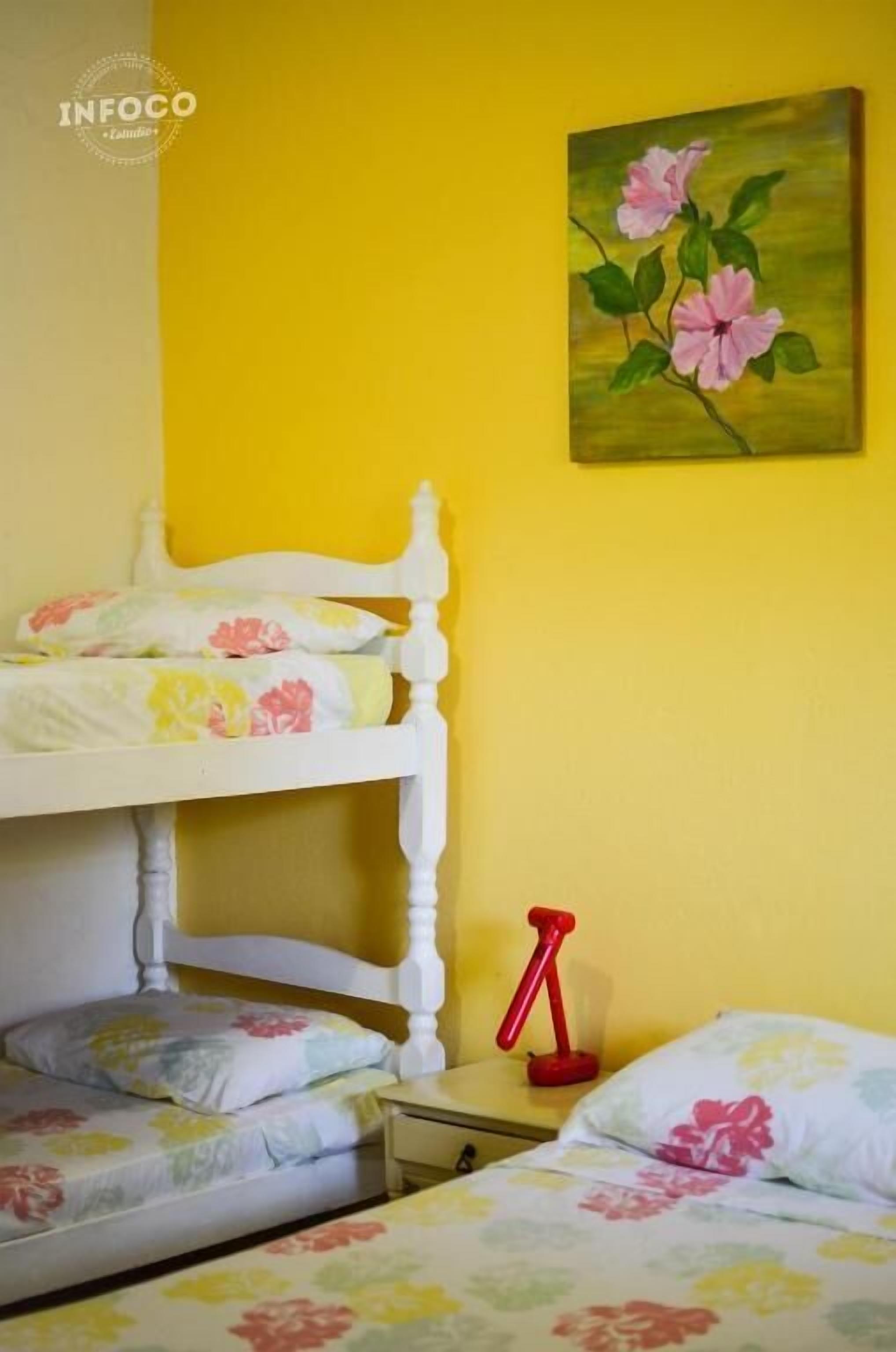 A room with a bunk bed, a bedside table, and a painting of flowers on the wall.