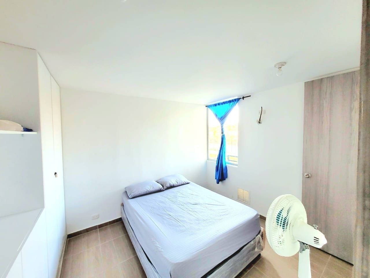 Une chambre à coucher avec un lit, une fenêtre avec des rideaux bleus, un ventilateur sur pied et une armoire.