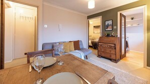 Ein Essbereich mit einem Holztisch, Stühlen und einem Weinglas. Im Hintergrund befindet sich eine Couch und eine Holzkommode.