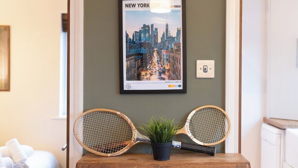 Ein gerahmtes Stadtbildposter von New York City an einer Wand, zwei Tennisschläger auf einem Holztisch und ein Blumentopf.