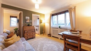 Ein gemütliches Wohnzimmer mit einer Couch, einer Holzkommode, einem Esstisch mit Stühlen, einer Lampe und einem Fenster mit Vorhängen.