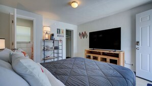 Quarto com uma cama grande, uma TV sobre um suporte de madeira, um guarda-roupa, uma prateleira com objetos e uma porta.