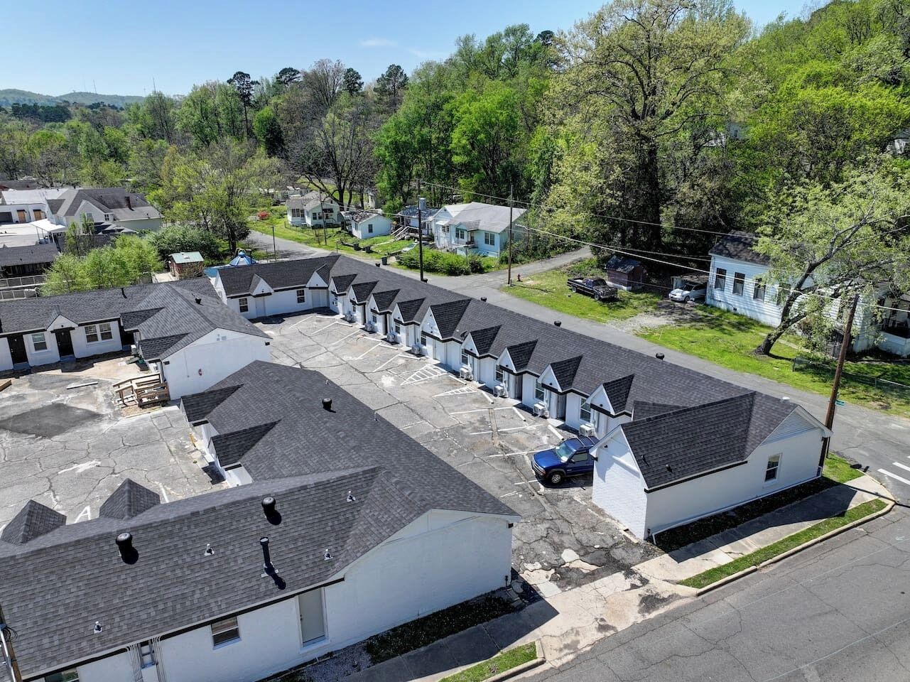 A residential area with multiple single-story houses, each with a dark roof and white walls. There are visible driveways and a few parked cars.