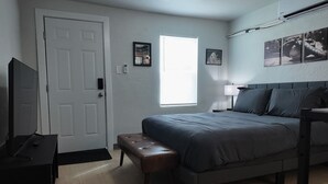 A bedroom with a bed, a bench, a TV, a door, and a window. - The Bambino at PWC (Hot Springs)