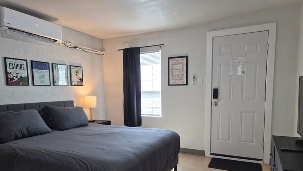 A bedroom with a bed, a nightstand with a lamp, a door, and a window with curtains. - The Boardwalk at PWC (Hot Springs)