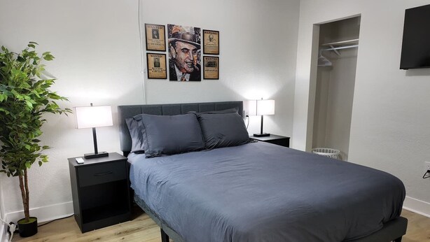 A bedroom with a bed, two bedside tables, a lamp, a potted plant, and a wardrobe. - The Capone at PWC (Hot Springs)
