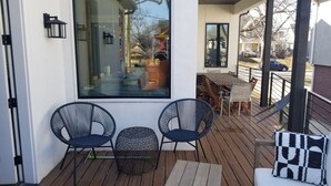 En träveranda med två stolar och ett litet bord, ett stort fönster som speglar en byggnad och ett räcke.