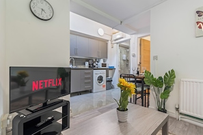 A modern kitchen with a washing machine, a dining area with a table and chairs, and a TV displaying 'NETFLIX'. - Private and Beautiful 1BR Apartment near South Park,Churchill Hospital, Oxford. (Oxford)