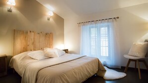 Un dormitorio con cabecera de madera, cama con ropa de cama blanca, mesita de noche pequeña, una silla con cojín y una ventana con cortinas translúcidas.