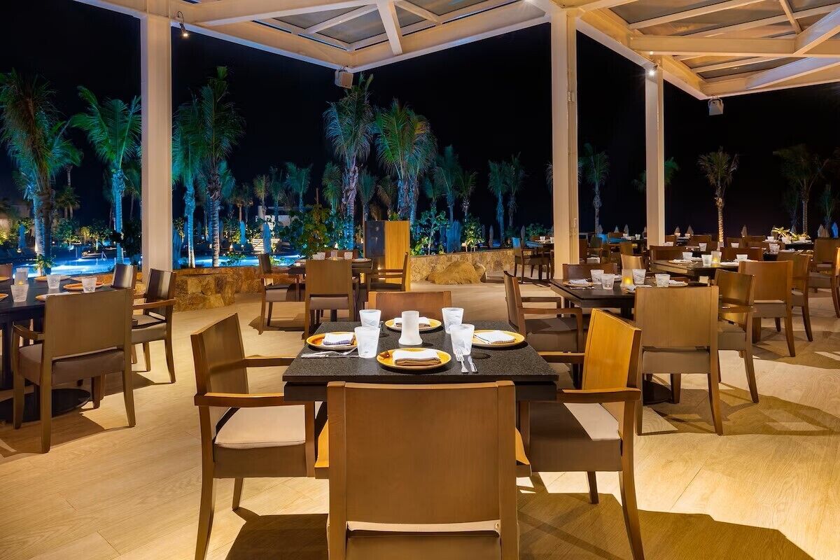 An outdoor dining area with tables and chairs, palm trees, and a pool in the background.