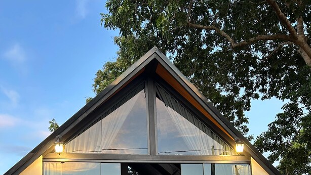 Una cabaña moderna con paredes de vidrio, una terraza de madera y una cama en su interior.