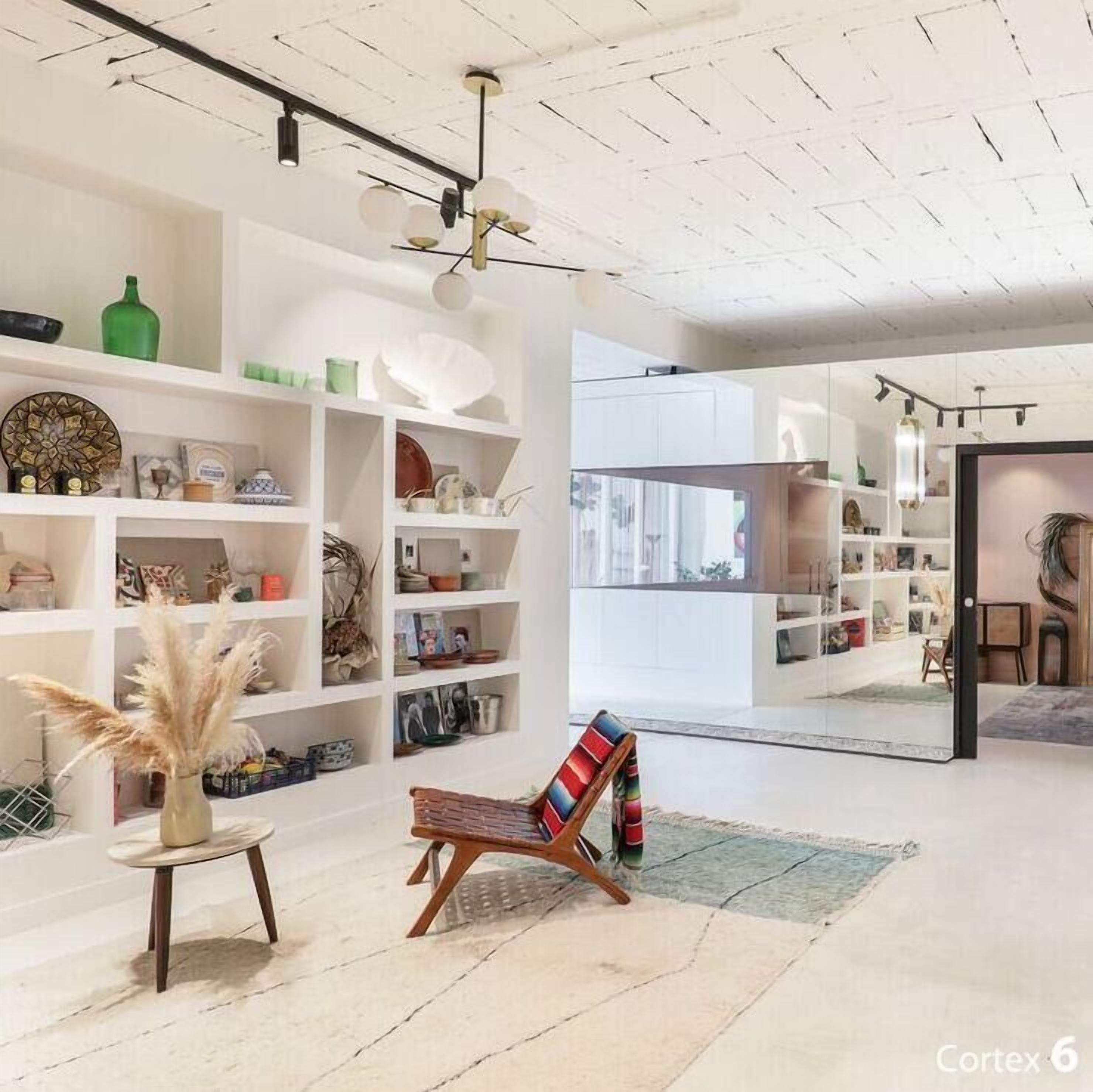 Een ruime kamer met witte muren en een hoge nok, met een zithoek in het midden bestaande uit een kleurrijke stoel en een klein tafeltje. Planken langs de muren tonen diverse decoratieve objecten en boeken.