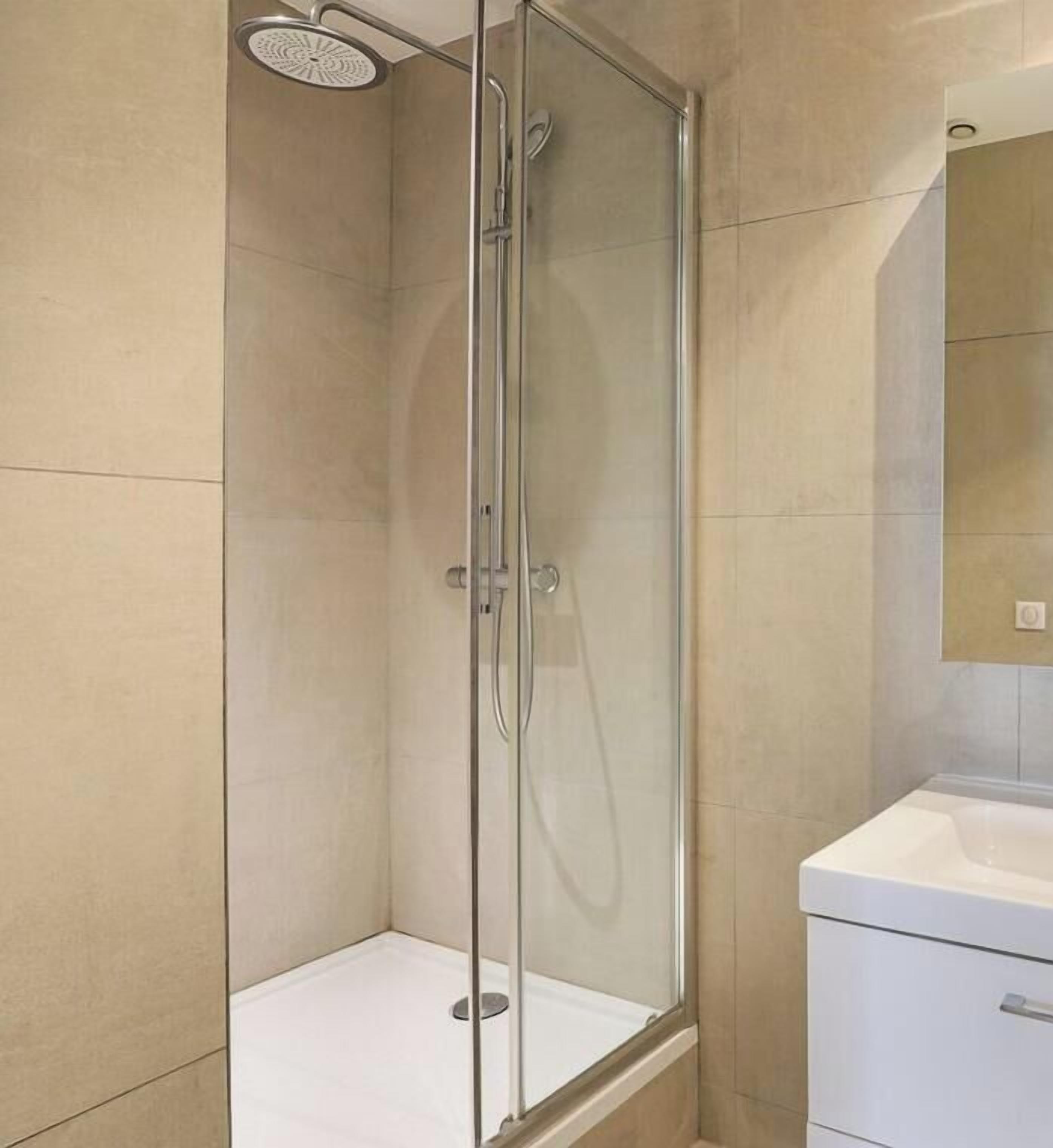 Une cabine de douche fermée par des parois en verre, dotée d’un pommeau de douche à main, d’un lavabo blanc et d’un miroir au-dessus du lavabo.