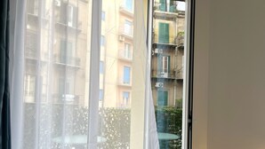 Vista a través de una ventana con cortinas translúcidas, mostrando un balcón con mesa y sillas, y un edificio con balcones y ventanas al fondo.