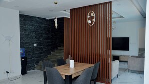 Un salon moderne avec une table à manger en bois, des chaises grises, un téléviseur noir et un escalier adossé à un mur foncé.