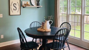 A dining area with a round table, four chairs, a chandelier, wall-mounted shelves, and a large window with a view of the outdoors. - Masters Rental 4-bedroom house in Golf Course Community (Evans)