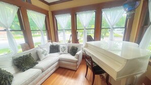 Una habitación con un piano blanco, un sofá seccional color beige con cojines decorativos y ventanas grandes con cortinas translúcidas.