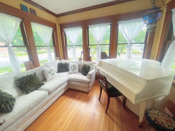 Una habitación con un piano blanco, un sofá seccional color beige con cojines decorativos y ventanas grandes con cortinas translúcidas.