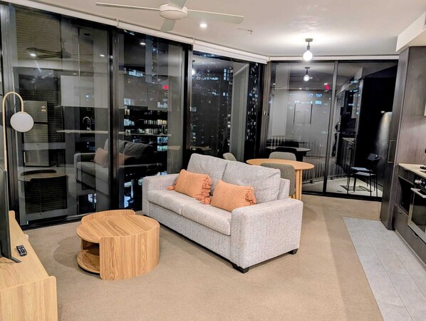 Una sala de estar moderna con un sofá gris, una mesa de centro redonda de madera y un separador de vidrio con vistas a la ciudad.