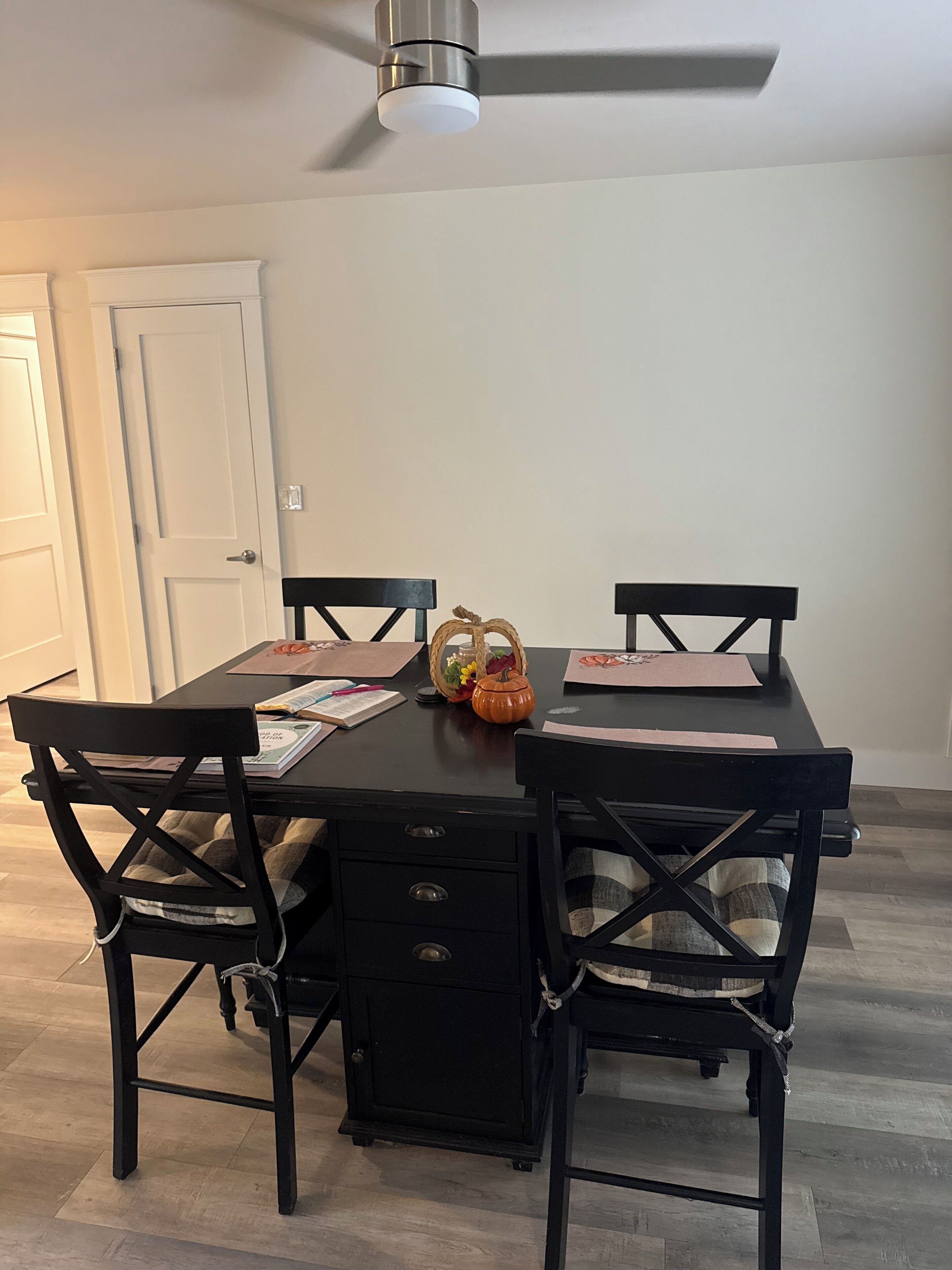 A dining table with four chairs, a ceiling fan, and a white door in the background.