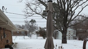 Une table de pique-nique recouverte de neige, avec un parasol, une maison en briques et une cour clôturée.