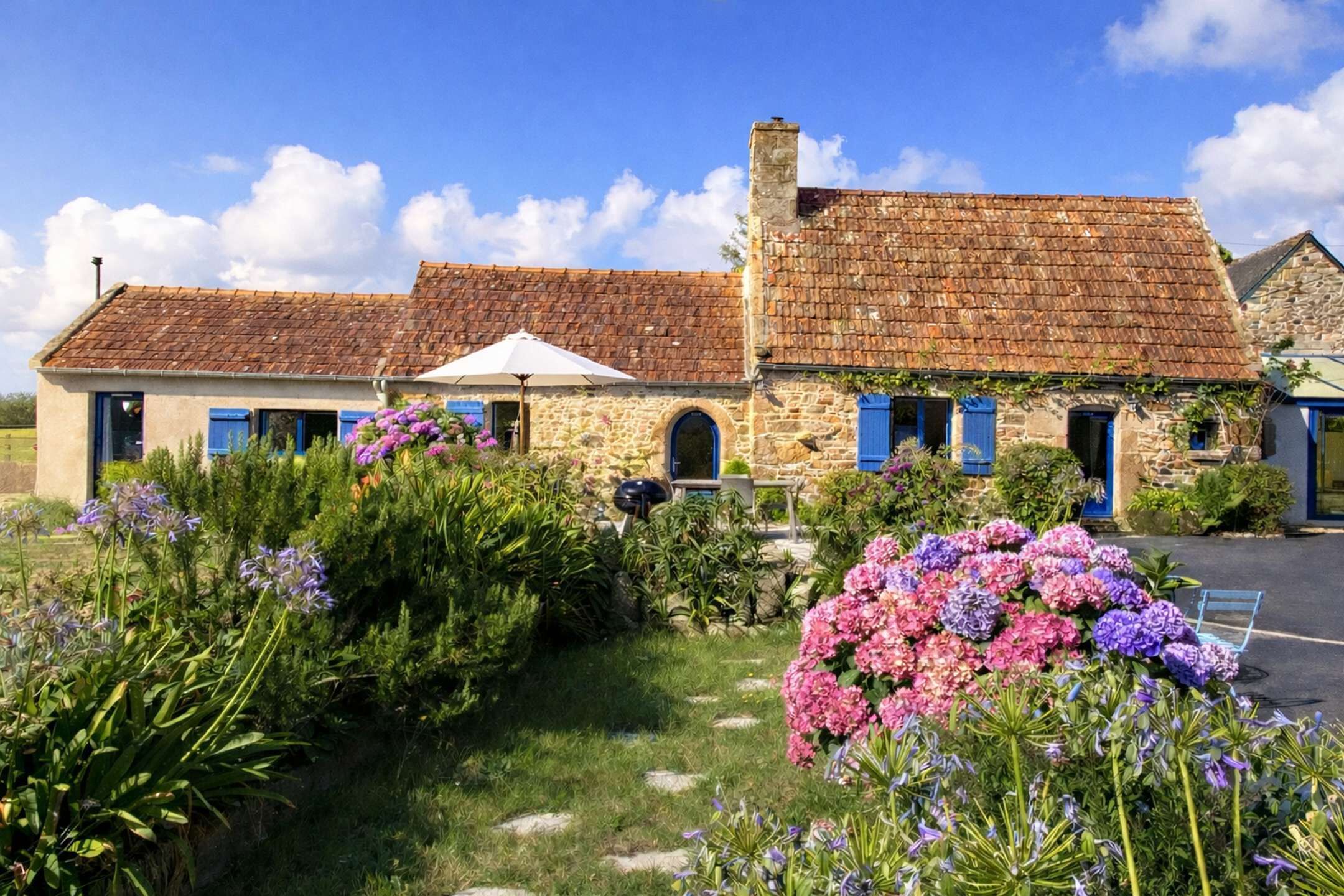 Un bâtiment en pierre avec des volets bleus, un jardin fleuri et un ciel dégagé.