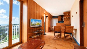 Ein modernes Interieur mit Holzboden, einem Flachbildfernseher, einem Essbereich mit Stühlen und Blick durch ein großes Fenster.