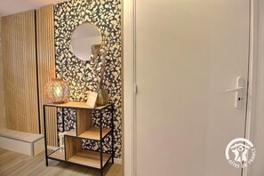 Ein Zimmer mit einer Wand aus Holzlatten, einem runden Spiegel, einem kleinen Holztisch mit Lampe und einer weißen Tür.