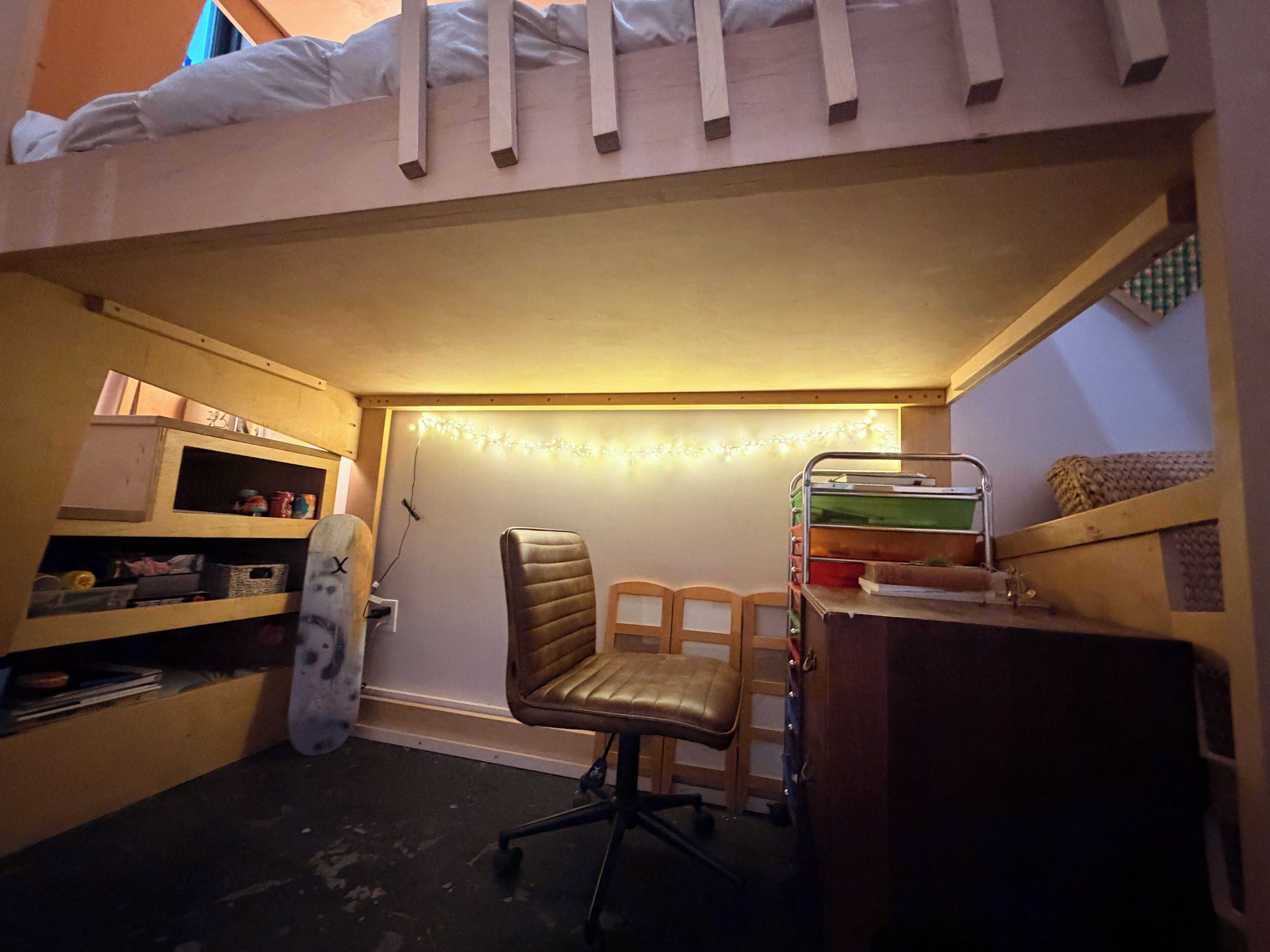 Una cama litera con escritorio y silla debajo, un estante con varios objetos, y una guirnalda de luces en la pared.