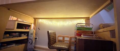 Una cama litera con escritorio y silla debajo, un estante con varios objetos, y una guirnalda de luces en la pared.