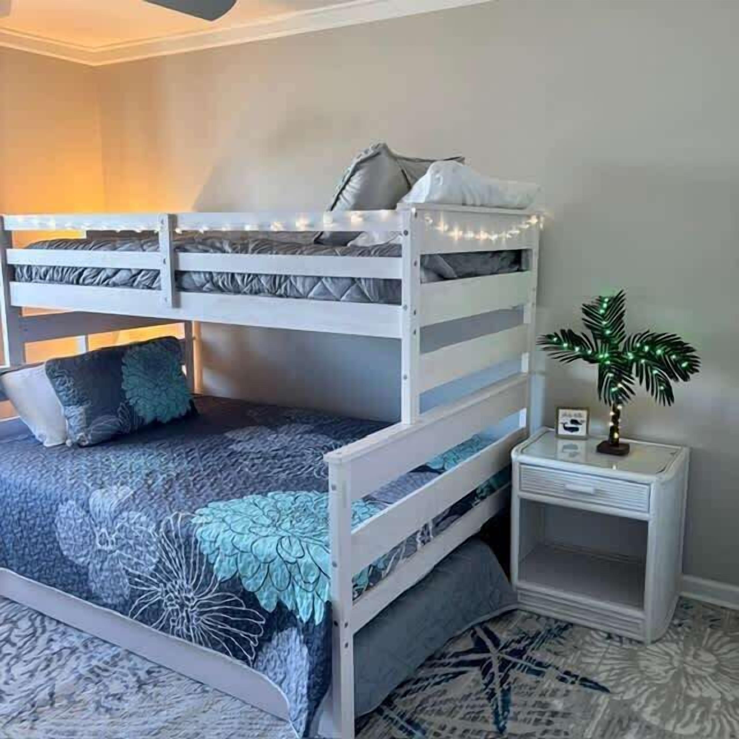 Uma cama beliche com colcha estampada de flores, uma mesinha de cabeceira com planta e um pequeno porta-retrato.
