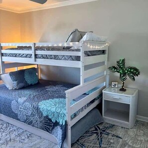 Uma cama beliche com colcha estampada de flores, uma mesinha de cabeceira com planta e um pequeno porta-retrato.