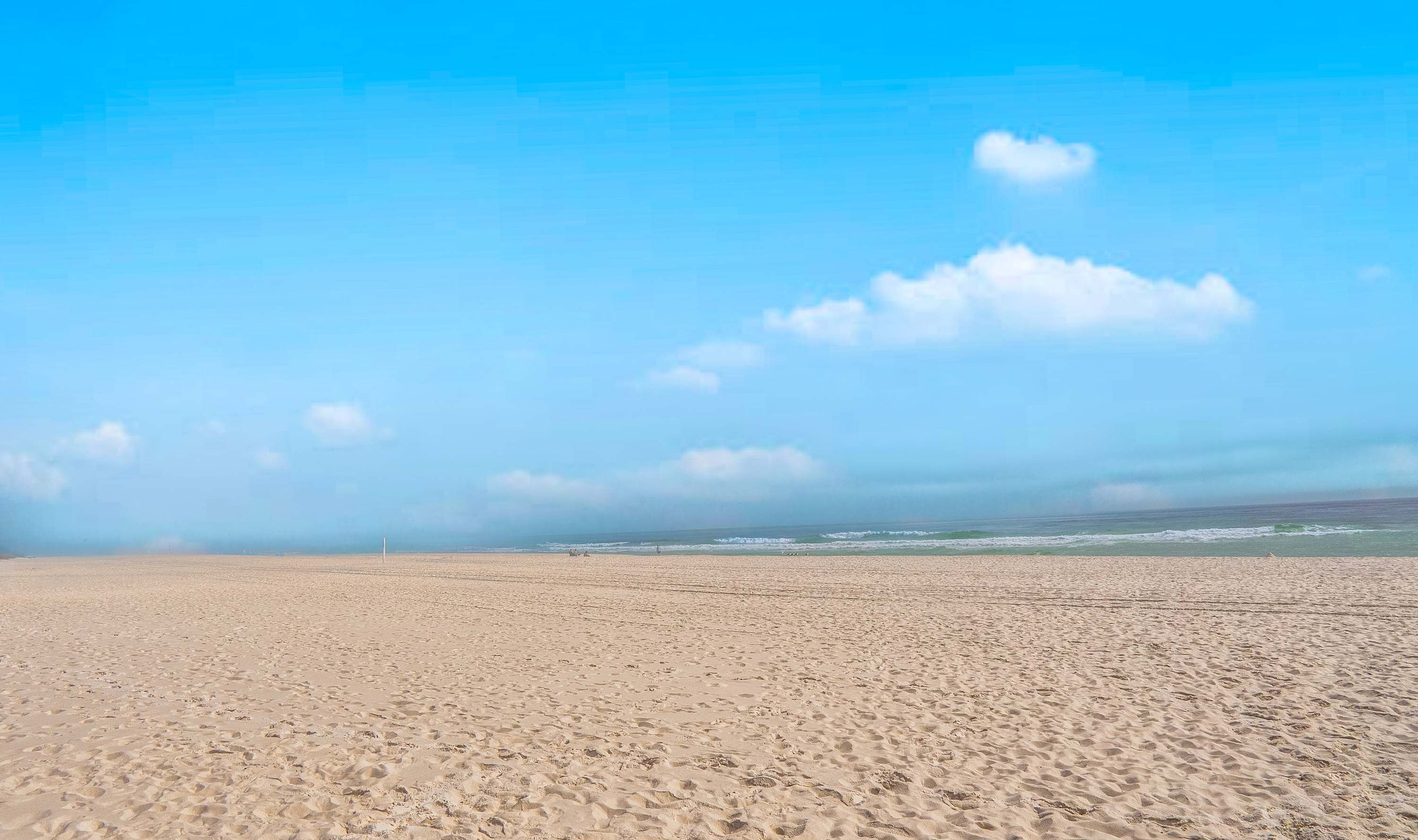A wide sandy beach with a clear blue sky and a few clouds.
