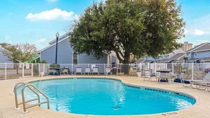 Una piscina con una escalera metálica, rodeada por una valla blanca, y un árbol al fondo.